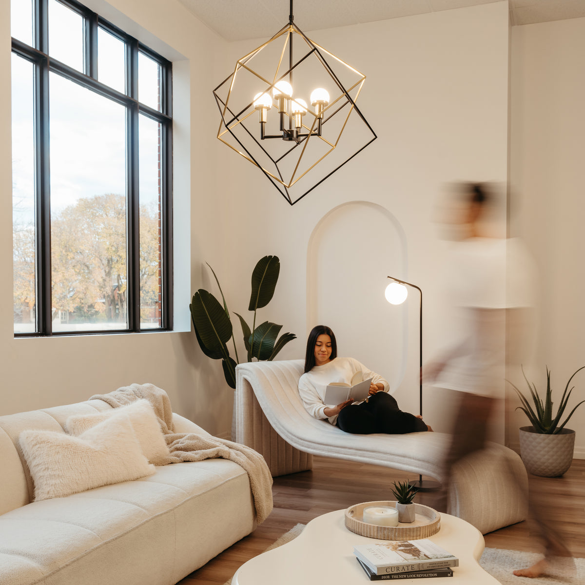 A modern, cozy living room with a woman reading on a curved chaise lounge, a blurred person walking by, soft beige furniture, large window, geometric chandelier, and potted plants. Warm natural light fills the space.