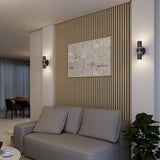 Modern living room with a grey sofa, a wooden slatted accent wall, abstract artwork, two wall sconces, and a leafy green plant in the corner. The space is softly lit and opens to a dining area in the background.
