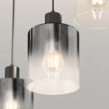 A close-up of a modern pendant light with a clear cylindrical glass shade and an exposed glowing filament bulb inside, suspended by a dark cord against a neutral background.