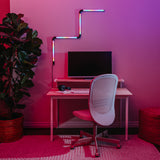 A modern desk setup with a computer monitor, keyboard, headphones, and a white mesh chair. Colorful LED lights are mounted on the wall above, and a large potted plant is nearby, all under pink and purple lighting.