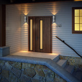 A modern wooden front door with vertical frosted glass panels is illuminated by wall lights. The entrance has stone steps, a metal handrail, and a house number 845 beside the door on light-colored siding.