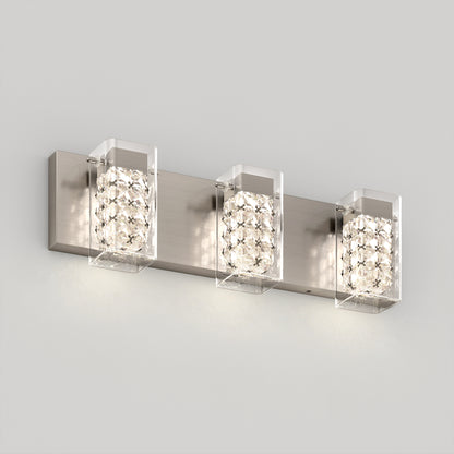 A modern wall-mounted vanity light fixture with three square lights, each featuring a metal base and crystal-like glass covers, emitting a soft white glow against a light gray wall.