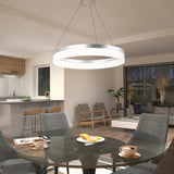 A modern dining area with a round glass table set for breakfast, gray upholstered chairs, a glowing circular pendant light above, and an open view of a cozy living room and kitchen in the background.