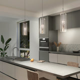 Modern kitchen with gray cabinets, built-in appliances, a large island with a sink, barstool seating, and three hanging pendant lights. A window shows greenery outside; decor includes plants and kitchen utensils.