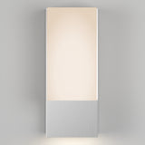 A modern wall sconce with a rectangular frosted glass shade and a brushed metal base, mounted on a light gray wall, emits soft, warm light.
