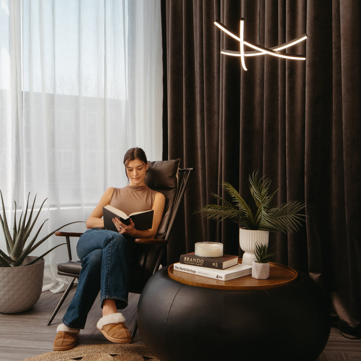 A woman sits in a chair reading a book in a cozy, modern living room with large windows, dark curtains, potted plants, and a round coffee table with books and a candle.