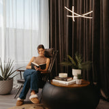 A woman sits in a chair reading a book in a cozy, modern living room with large windows, dark curtains, potted plants, and a round coffee table with books and a candle.