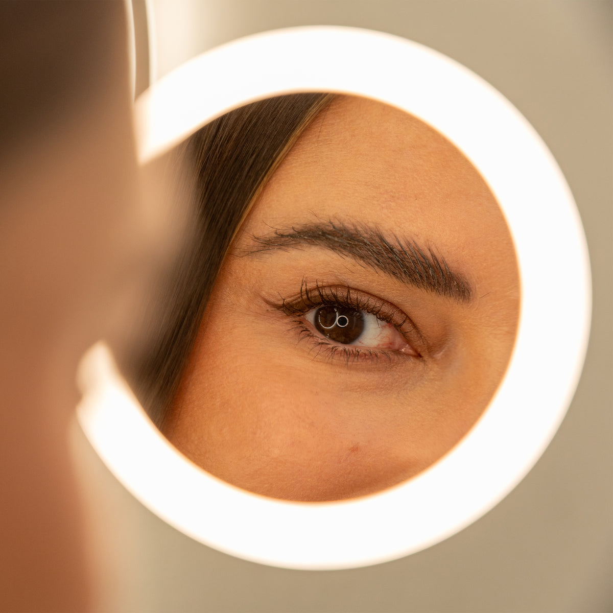 A close-up of a womans eye reflected in a circular mirror with a ring light, highlighting her eyebrow, eyelashes, and smooth skin.