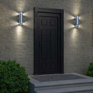 A black door set in a brick wall is illuminated by two Artika C7 indoor-outdoor wall lights on either side, while two green bushes frame the steps leading up to the entrance.