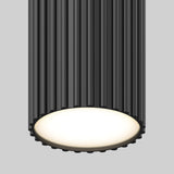 A close-up of a modern, cylindrical black ceiling light fixture with vertical grooves and a glowing white circular light at the bottom, set against a plain gray background.