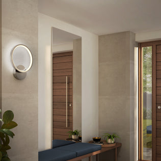 A modern entryway with beige walls, an Artika Solis modern LED indoor-outdoor wall light 5 CCT for accent lighting, a wooden bench with blue cushion, potted plants, and large wooden doors. Tall windows fill the space with natural light—ideal for contemporary homes.