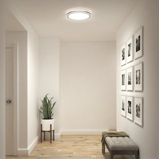 A minimalist hallway with light wood floors, white walls, a potted plant on a stand, a bench with a throw blanket, black-and-white photo frames, and the Artika Altitude 15 in. LED modern flush mount light 5 CCT on the ceiling.