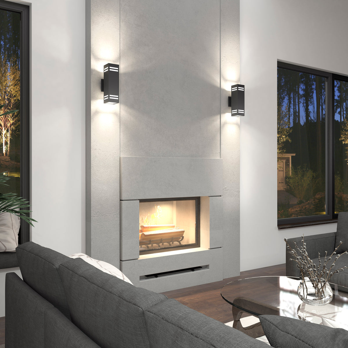 Modern living room with a minimalist gray fireplace, two wall sconces, gray sectional sofas, a glass coffee table, and large windows overlooking a wooded outdoor scene at dusk.