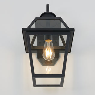The Artika Colonial outdoor lantern wall light is mounted on a white wall, showcasing a visible glowing filament bulb inside its rectangular black metal and glass enclosure.