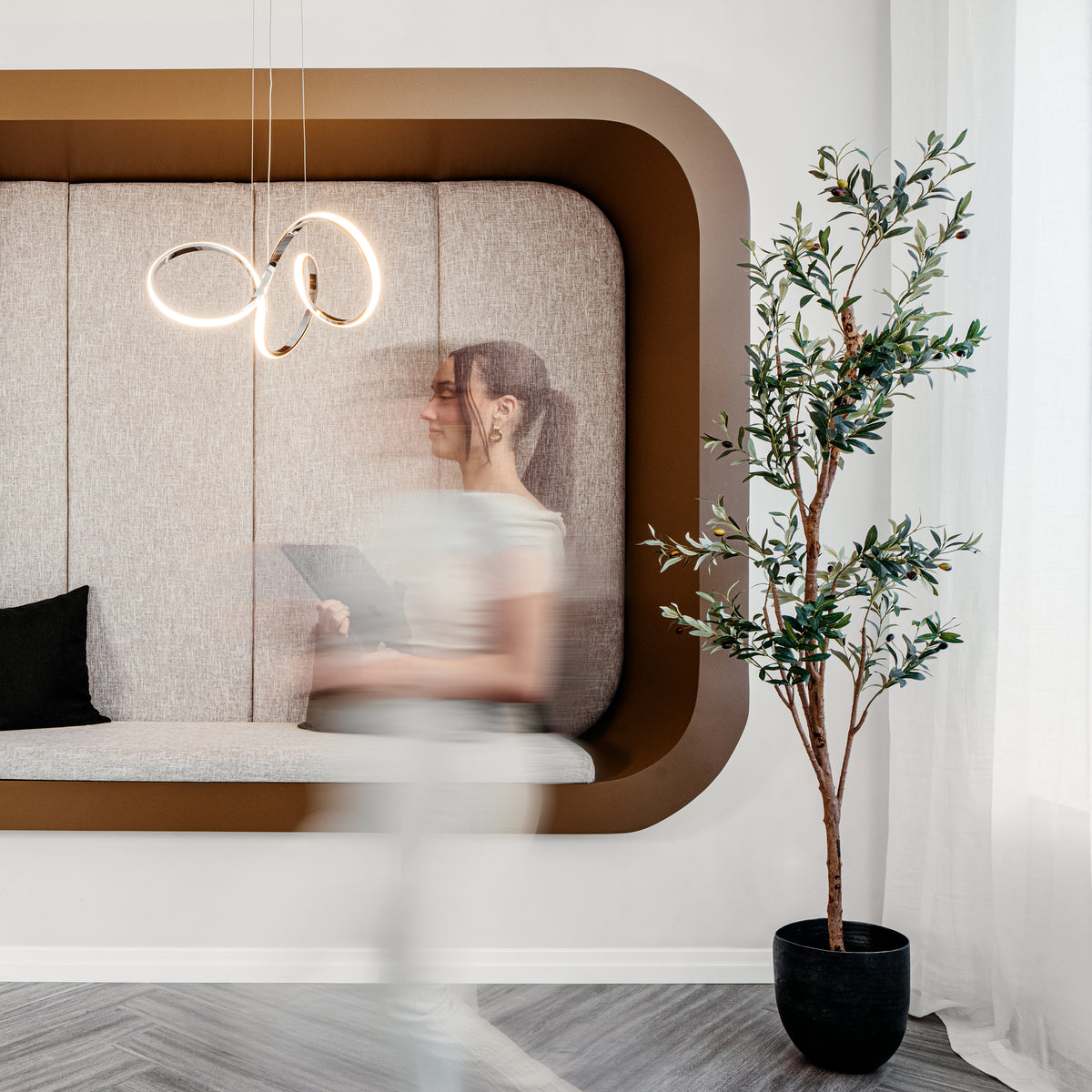 A modern lounge features a curved beige wall seat, black cushion, potted plant, and the Swirl LED pendant light by Artika. White curtains let in natural light from the right as a woman passes by, her figure blurred from motion.