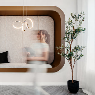 A modern lounge features a curved beige wall seat, black cushion, potted plant, and the Swirl LED pendant light by Artika. White curtains let in natural light from the right as a woman passes by, her figure blurred from motion.