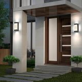 Modern house entrance with a wooden door, vertical glass panel, two wall lights, potted plants on each side, and stone steps leading to the door. The exterior features a mix of concrete and wood finishes.