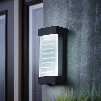 The Artika Percy LED indoor-outdoor wall light, featuring a modern black finish with frosted glass and horizontal lines, is mounted on a textured gray wall near a window and door, softly illuminating the area.