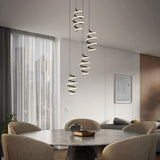 A modern dining room with a round table and four beige chairs, featuring three spiral pendant lights hanging above. Large window with sheer curtains and city view, minimalist decor, and a wooden accent wall.