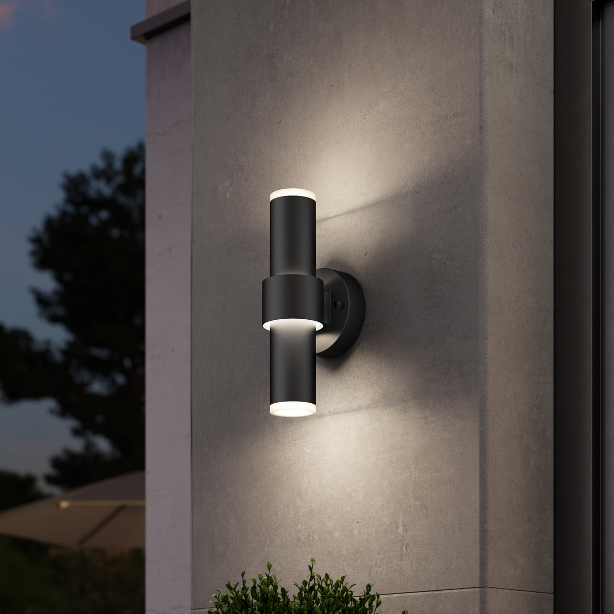 A modern black cylindrical outdoor wall light fixture mounted on a gray concrete wall, illuminating upwards and downwards in the evening. Trees and part of an umbrella are visible in the blurred background.