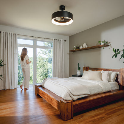 A woman stands by large windows in a bright bedroom with wooden floors and an Artika Austin 23 in. LED fandelier ceiling fan.