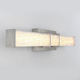 A modern wall sconce with a rectangular brushed metal frame and a textured, illuminated glass panel, mounted on a light gray wall.