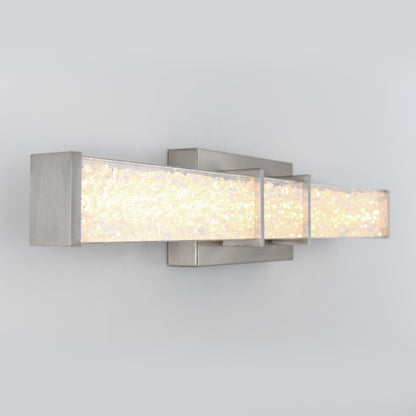 A modern wall sconce with a rectangular brushed metal frame and a textured, illuminated glass panel, mounted on a light gray wall.