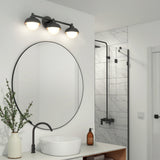 Modern bathroom with a round mirror, black fixtures, white vessel sink, marble-tiled shower, wooden vanity with shelves, potted plant in a striped vase, and soap dispenser on the counter. Three-bulb light fixture above mirror.