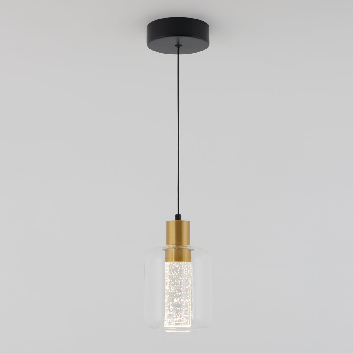 A modern pendant light with a black ceiling mount, a black cord, a gold fixture, and a cylindrical clear glass shade surrounding a textured inner diffuser.