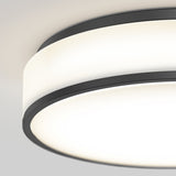 A close-up view of a modern, round ceiling light fixture with a black metal frame and frosted glass emitting a soft, bright glow against a white ceiling.