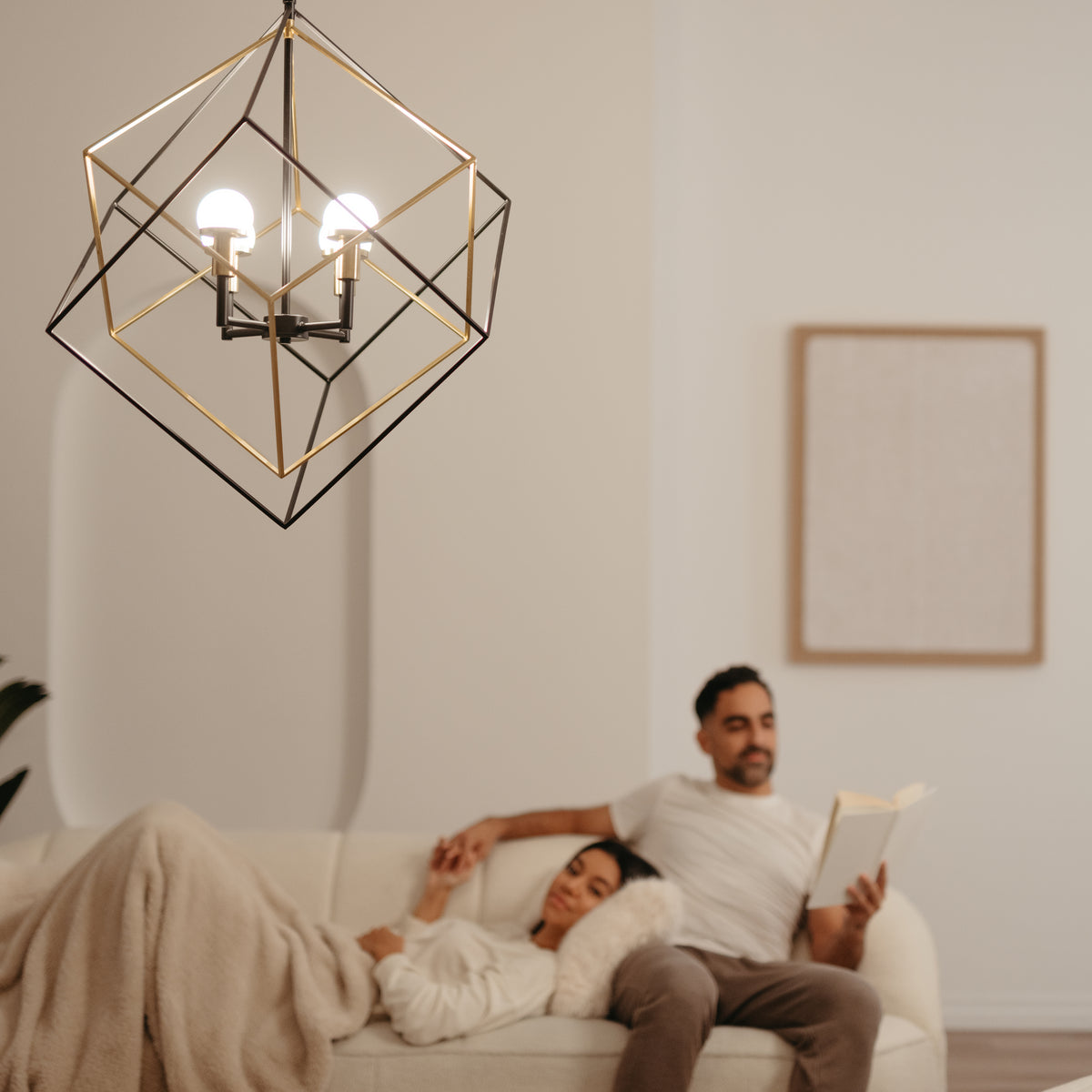 A modern geometric light fixture hangs in the foreground while a woman rests on a sofa under a blanket and a man sits beside her reading a book in a softly lit, minimalist living room.