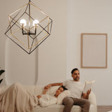 A modern geometric light fixture hangs in the foreground while a woman rests on a sofa under a blanket and a man sits beside her reading a book in a softly lit, minimalist living room.