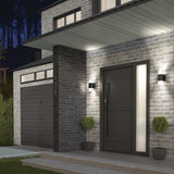 Modern home exterior at dusk with gray brick walls, black front door with a vertical glass panel, black garage door, two potted topiary plants by the entrance, and wall-mounted lights illuminating the porch.