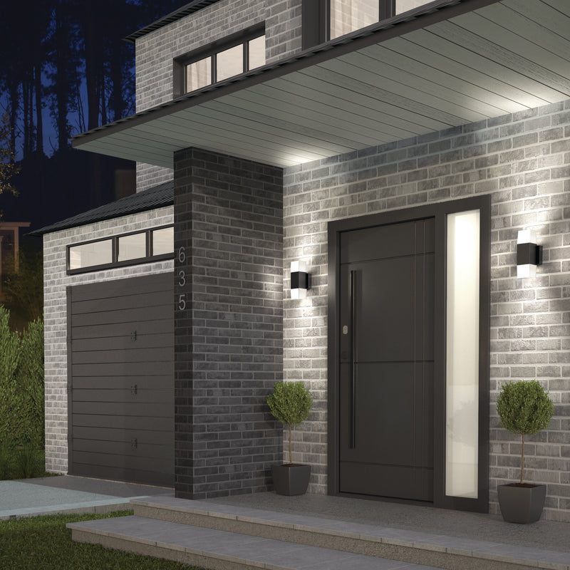 Modern house exterior with gray brick walls, dark gray garage and front doors, tall window, two potted plants, and Artika Lennox indoor-outdoor LED wall lights (3 CCT) illuminated at dusk. House number 635 is visible.