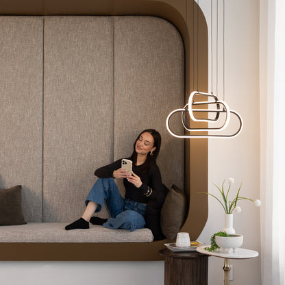 A woman sits on a cushioned bench, smiling at her phone. Artika’s Lewis modern LED pendant light 3 CCT hangs nearby, while a small table with a vase, cup, and plate rests in the foreground.