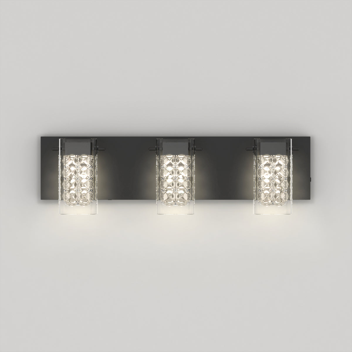 A modern wall sconce with three lights, each featuring a cylindrical glass shade and a black rectangular backplate, mounted on a light gray wall. The lights are lit, emitting a soft glow.