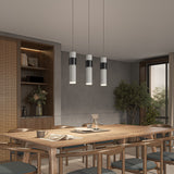 A modern dining room with a wooden table and chairs, pendant lights above, built-in shelves with decor items and books, a vase with flowers, and a large potted tree next to a window.