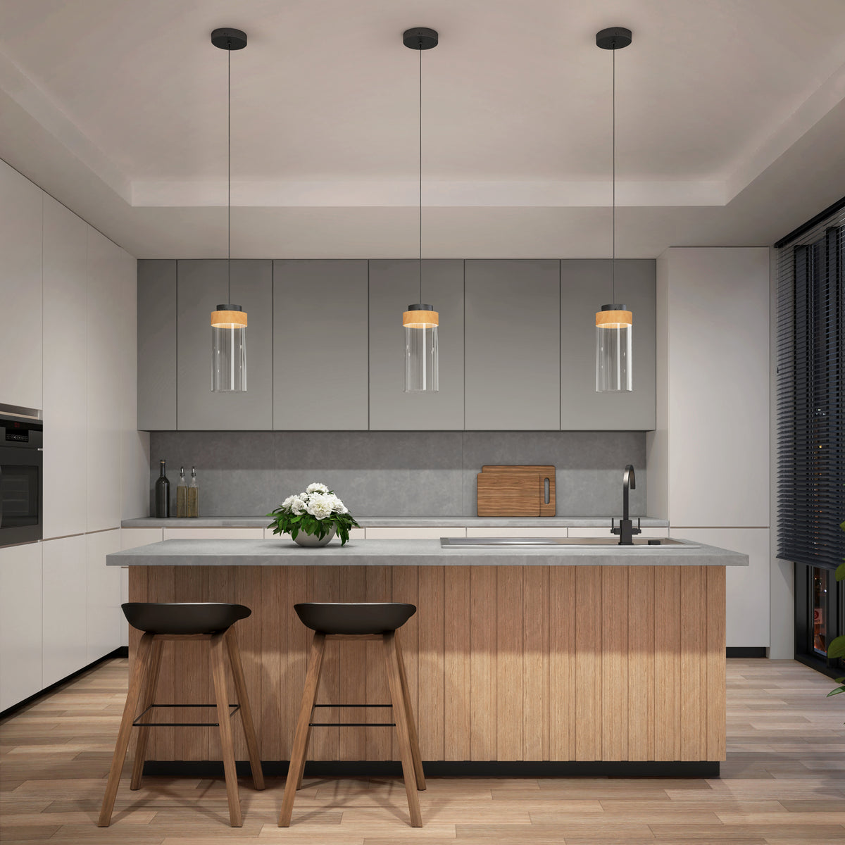 Modern kitchen with a wooden island, two bar stools, and three Artika Hanson 1-light LED pendant lights overhead. White and gray cabinets, built-in oven, vase with white flowers on the island, and dark window blinds.