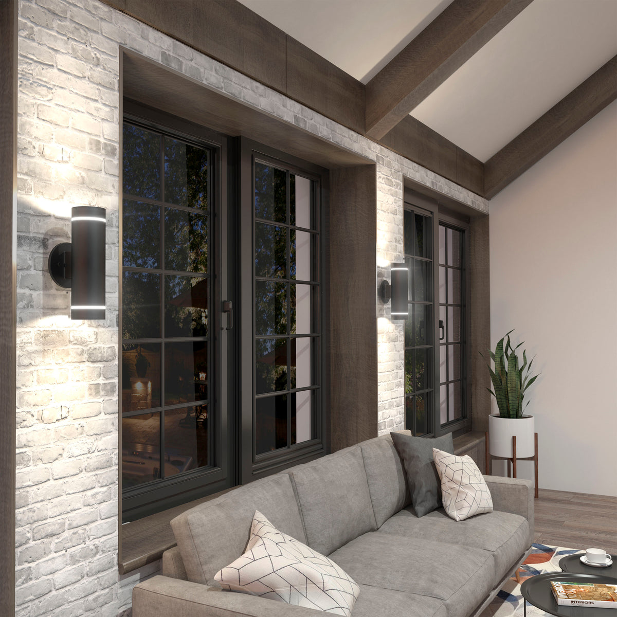 A modern living room with a gray sectional sofa, geometric-patterned pillows, large windows with black frames, brick accent walls, two Artika C7 indoor-outdoor wall lights, and a potted plant in the corner.