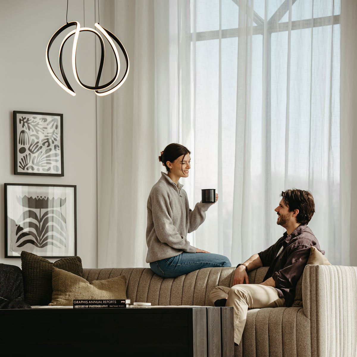 A woman sits on the back of a beige sofa holding a mug, smiling at a man seated on the sofa. Modern art hangs on the wall, and a unique pendant light is above them in a bright, cozy living room.