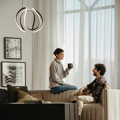 A woman with a mug smiles at a man on the couch in a modern, sunlit living room featuring framed art, stacked books, and the Artika Vivaldi Big LED pendant light (5 CCT) overhead.
