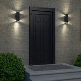 A modern black door with decorative panels is centered on a light brick wall, flanked by two lit wall sconces. Green bushes frame the doorway, and three steps lead up to the entrance.