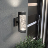 A modern outdoor wall sconce with a cylindrical, textured glass shade glows warmly, mounted on a gray wall near black horizontal-paneled doors, with green shrubbery below.