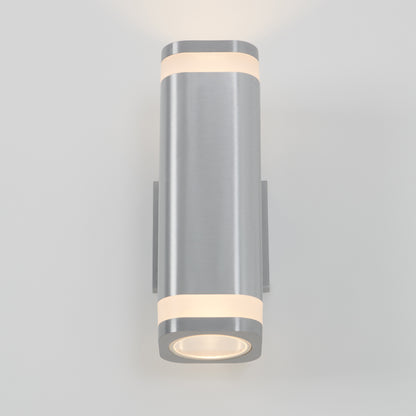 The Artika Mettle LED indoor-outdoor wall light features a modern cylindrical design in brushed silver with rounded edges, casting soft white light both upward and downward on a plain, light-colored wall.