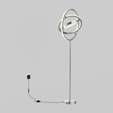A modern floor lamp with circular, intersecting LED rings on a tall metal stand, plugged into a wall outlet with a visible power cord against a plain gray background.