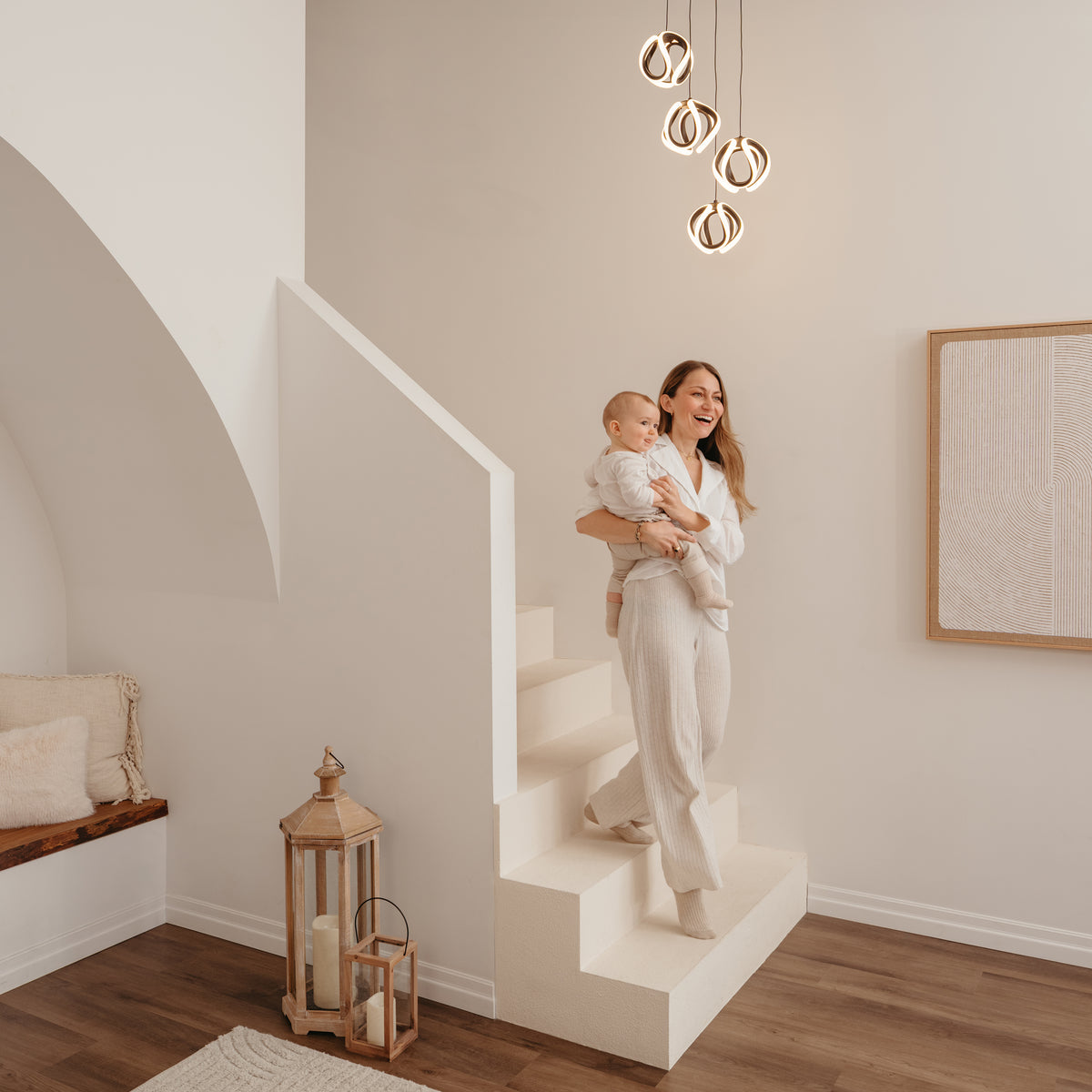 A woman in light clothing holds a baby on modern white stairs in a stylish living space with wood floors, neutral decor, and the Artika Vivaldi modern LED 4-light pendant (5 CCT) providing warm lighting.