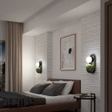 A modern bedroom with a brown bed, gray and white bedding, light brick walls, abstract art above the headboard, and two wall sconces with round bulbs and small potted plants. A side table is next to the bed.