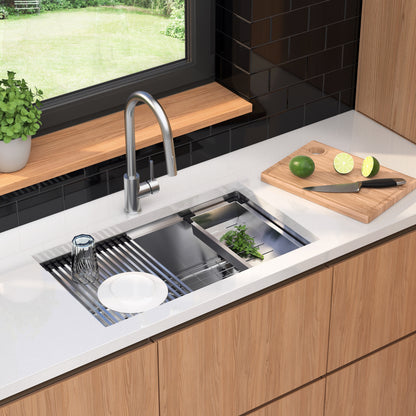 A modern kitchen features the Artika Newport 33 in. single bowl stainless steel workstation sink, a metal faucet, a drying rack with glass and plate, fresh herbs in the sink, and a wooden cutting board with knife, lime, and lemon slices on the counter.