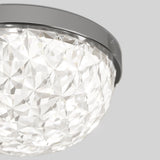 A close-up of a modern ceiling light fixture with a geometric, faceted glass cover and a sleek metal trim, emitting bright white light. The background is a plain light gray.