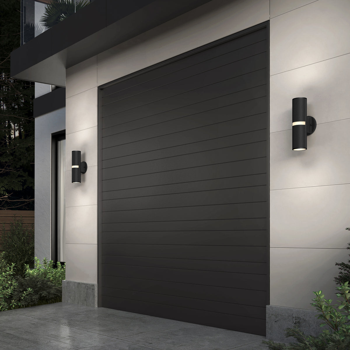 A modern house exterior features a large dark gray paneled garage door, flanked by two Artika Groove LED indoor-outdoor wall lights in sleek black. Green plants are near the base, with trees visible in the background.